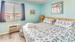 Ein Schlafzimmer mit Bett, Deckenventilator, Fenster mit Vorhängen, einem Wandbild und einer Nachttischlampe.