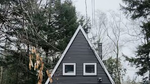 A small, dark-colored, triangular-shaped cabin with a front door, two windows on the upper floor, and a small porch. - Cozy & Modern A-Frame in the woods w/HOT TUB (Tamworth)