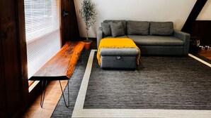 A cozy attic room with a grey sofa, a small ottoman, and a bench. - Cozy & Modern A-Frame in the woods w/HOT TUB (Tamworth)