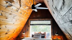 A cozy attic bedroom with a sloped ceiling, wooden walls, a bed with a 'Be Our Guest' pillow, a ceiling fan, and a window with a view. - Cozy & Modern A-Frame in the woods w/HOT TUB (Tamworth)