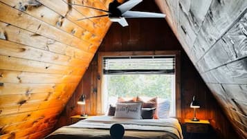 A cozy attic bedroom with a sloped ceiling, wooden walls, a bed with a 'Be Our Guest' pillow, a ceiling fan, and a window with a view.