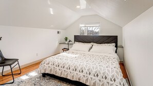 Ein Schlafzimmer mit geneigter Decke, einem Bett mit geometrischem Muster auf der Tagesdecke, einem Nachttisch mit Lampe, einem Sessel und einem Fenster mit Jalousien.