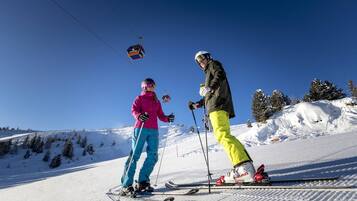 A ski slope with two skiers, a ski lift in the background, and snow-covered trees.