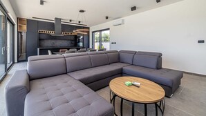 Una sala de estar moderna con un sofá seccional, una mesa de centro redonda y una cocina con armarios oscuros e iluminación colgante.