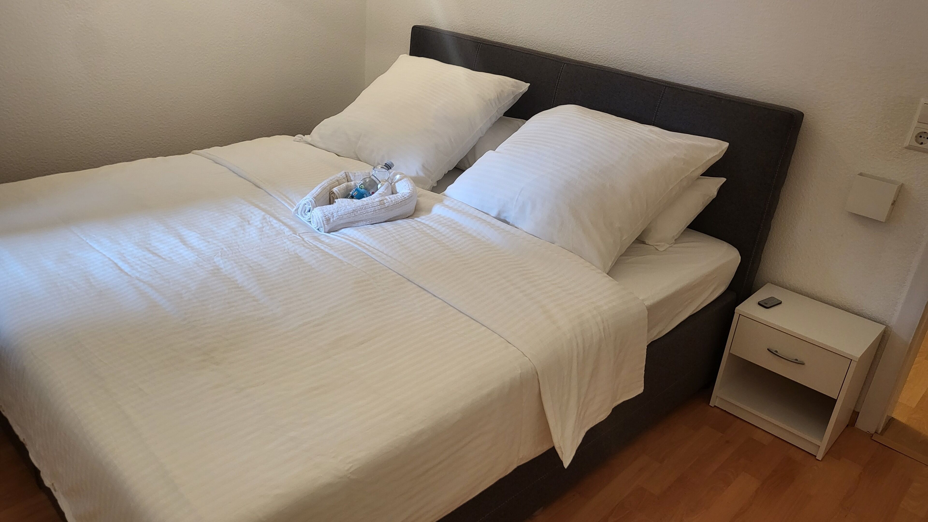 A neatly made bed with white linens and pillows, a small bedside table with a lamp, and a wall socket.