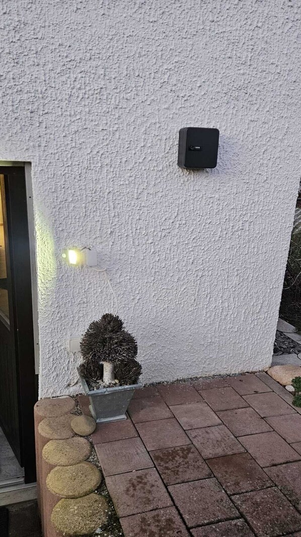 A white textured wall with a black intercom, a potted plant, and a light fixture.