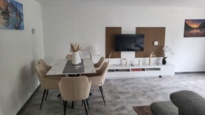 Un saló modern amb zona de menjador, amb taula de marbre, cadires beix, un moble blanc per a l'aparell d'entreteniment i un televisor penjat.