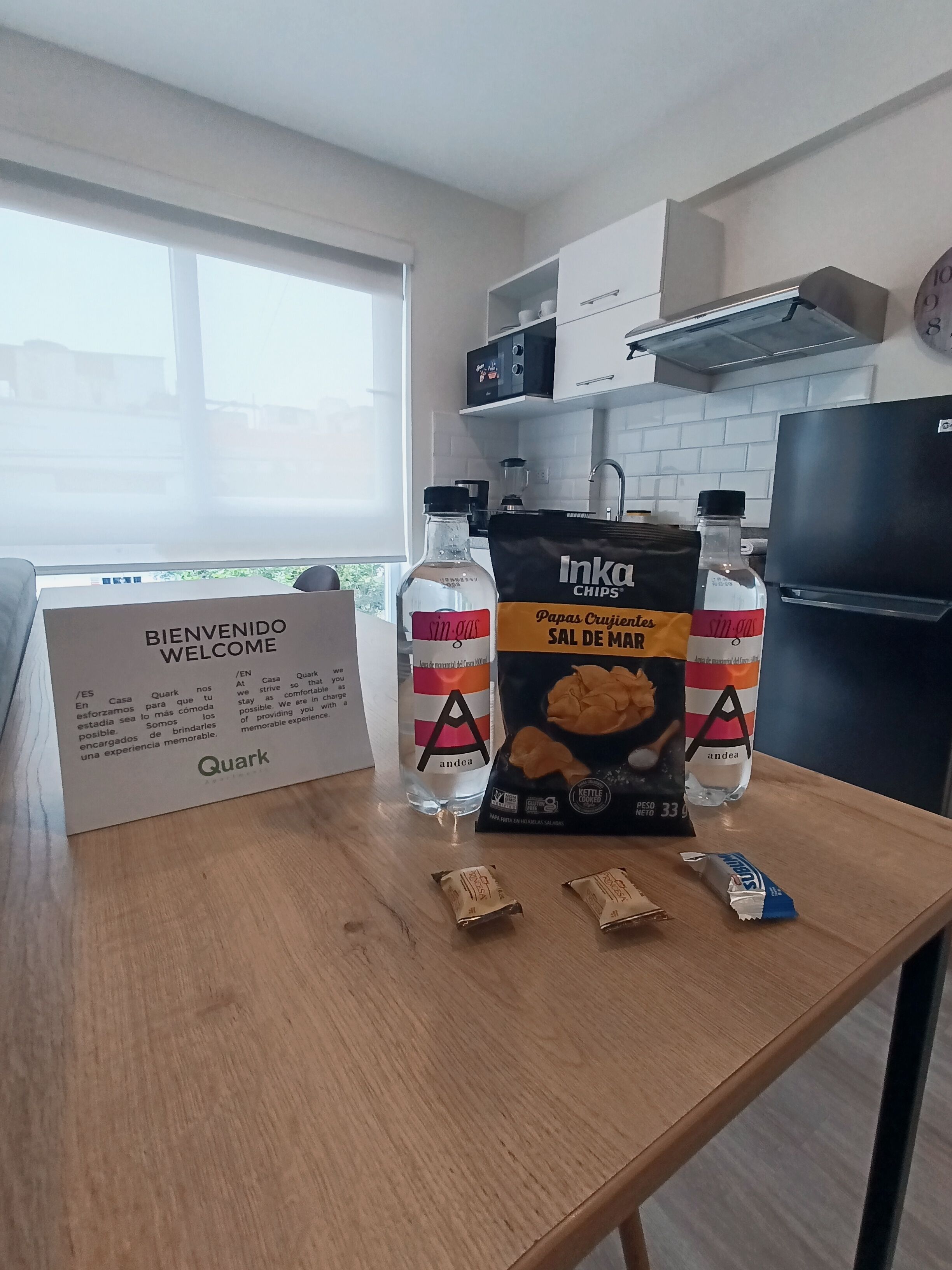 Una cocina con un cartel de bienvenida, un refrigerador, una ventana y una mesa con una bolsa de papas, una botella y dos paquetes.
