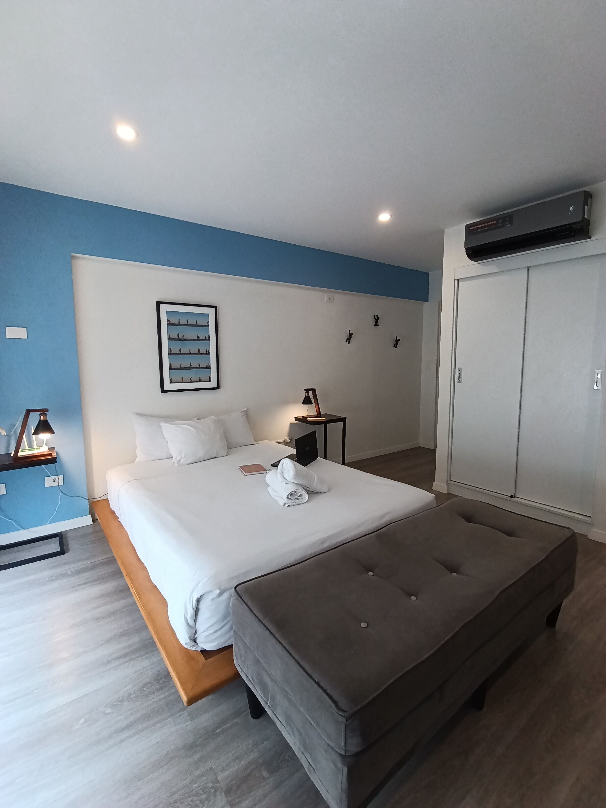 Un dormitorio con cama, mesita de noche con lámpara, armario y un cuadro en la pared.