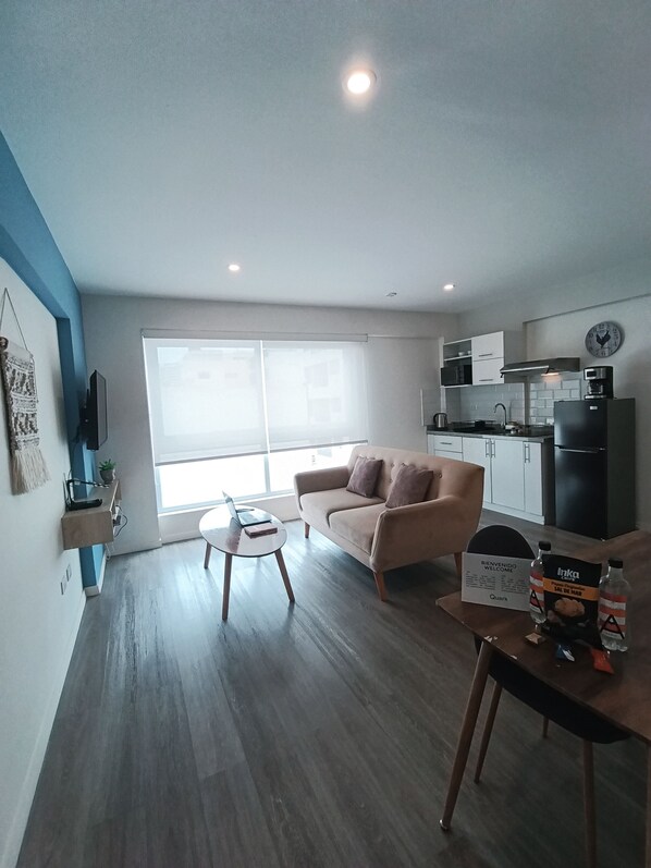 A modern apartment with a sofa, a small table, a kitchen area, and a large window. - Casa Quark Apartments Lima (Lima)