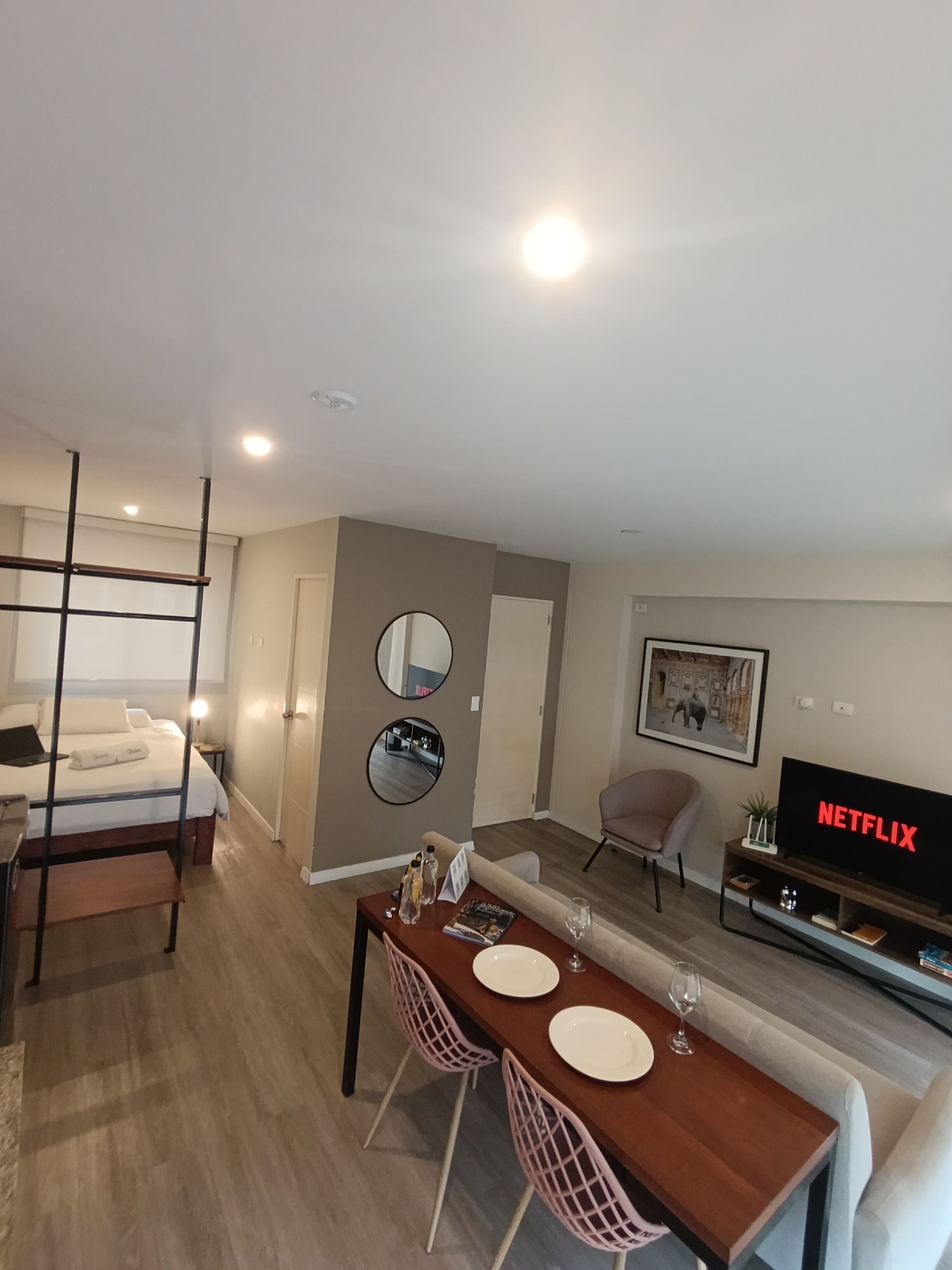 A modern hotel room with a bed, a dining area with a table and chairs, a TV displaying 'NETFLIX,' and a mirror on the wall.