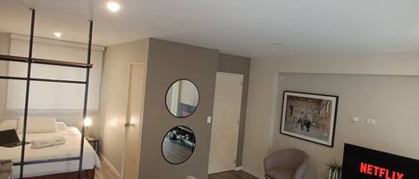 A modern hotel room with a bed, a dining area with a table and chairs, a TV displaying 'NETFLIX,' and a mirror on the wall.