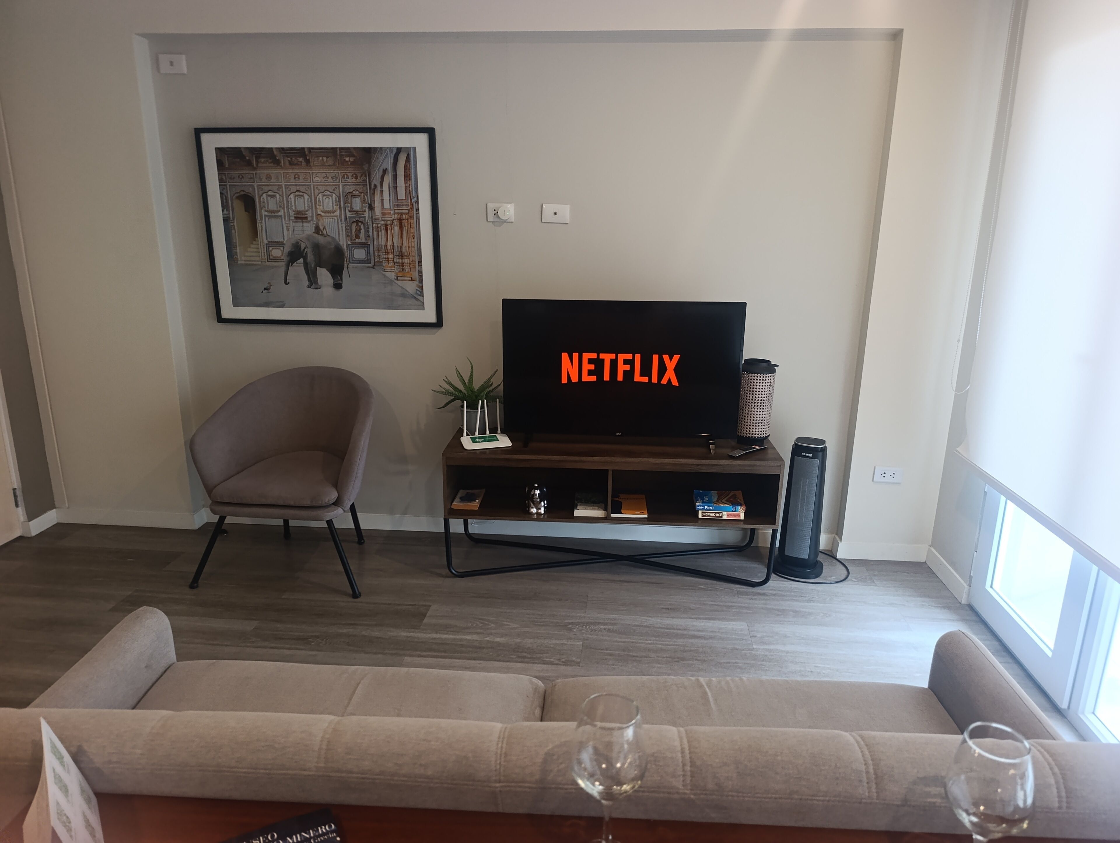 Una sala de estar moderna con un televisor de pantalla plana, un cuadro enmarcado, una silla, un sofá y una copa de vino.