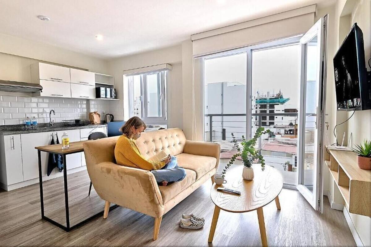 A modern apartment with a kitchen, a living area with a sofa, a dining table, and a large sliding glass door to a balcony.