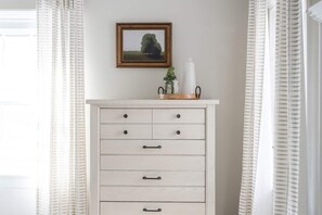A white dresser with drawers and knobs, a framed picture above it, and a small plant on top.