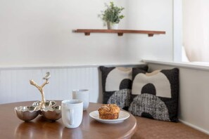 A wooden table with a decorative bird, a white mug, and a plate with a pastry. There are two white cups and a silver bowl. The background includes a white wall, a black and white patterned pillow, and a wooden shelf with a plant.