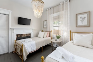 A bedroom with a fireplace, a television, a chandelier, a bed, a bedside table, and a window with curtains.