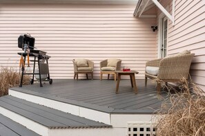 A covered outdoor deck with a grill, two wicker chairs, a table with drinks, and a wicker sofa.