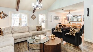 Un salon spacieux avec un grand canapé, un fauteuil noir et une table basse ronde. On y trouve également un coin repas avec une table et des chaises en bois. Le salon est doté de poutres apparentes au plafond et d'un lustre.