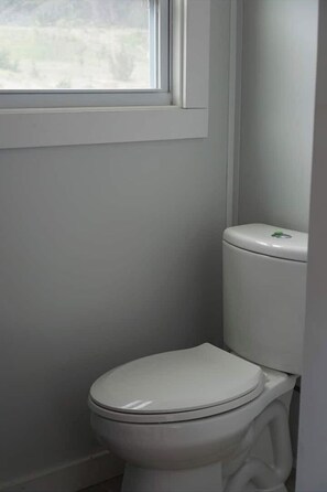 A white toilet with a green flush handle in a bathroom with white walls and a window above it. - Birch Tiny House Rental (Bandera)