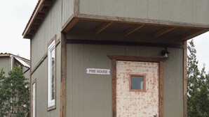 A small, single-story building with a wooden deck in front. A sign on the building reads "Pine House." - Pine Tiny House Rental (Bandera)