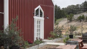 A red barn with a white door and windows, surrounded by picnic tables and barrels, with a gravel path and potted plants. - Pine Tiny House Rental (Bandera)