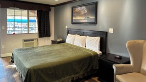 A hotel room with a bed, a chair, a nightstand, a painting, and a window with curtains. - Corona Inn (Corona)