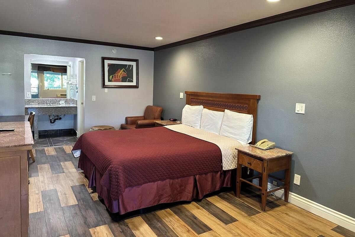 A bedroom with a large bed, a wooden nightstand, a chair, a desk, and a bathroom visible through an open door.