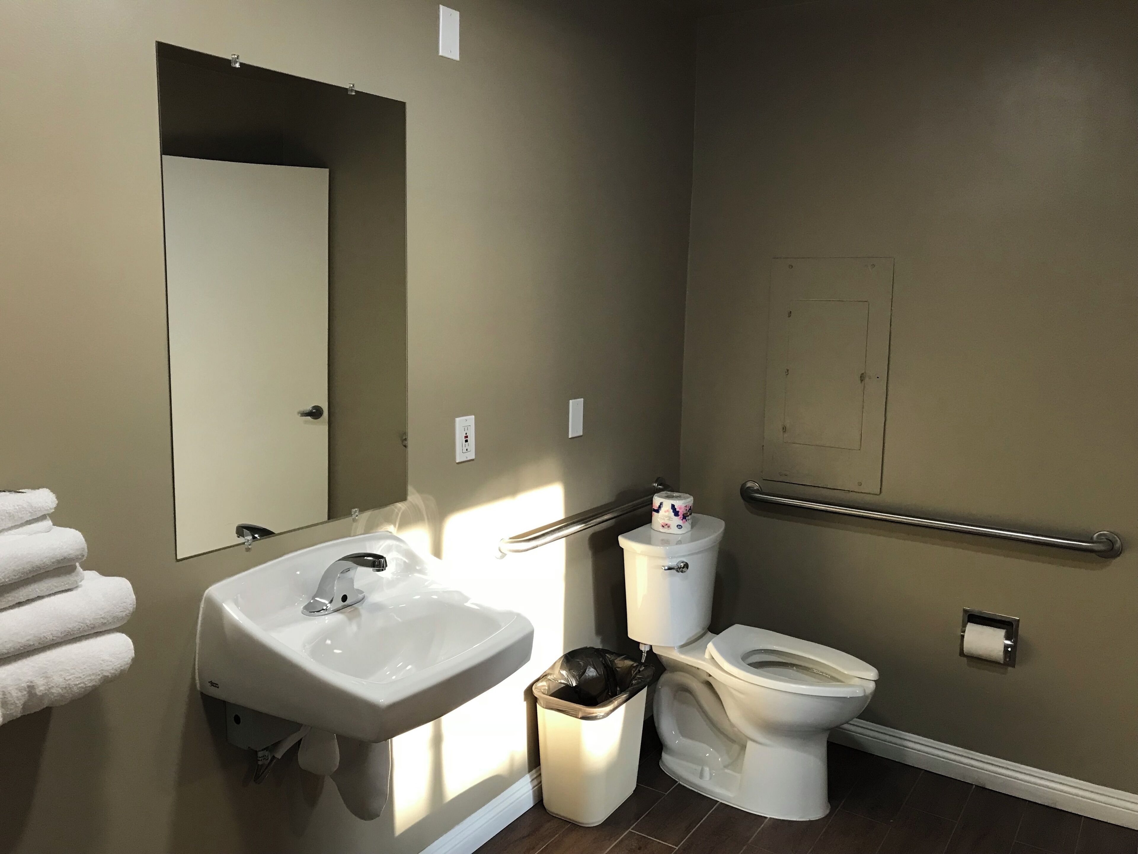 A bathroom with a sink, toilet, and mirror. There is a trash can and a towel rack with towels.
