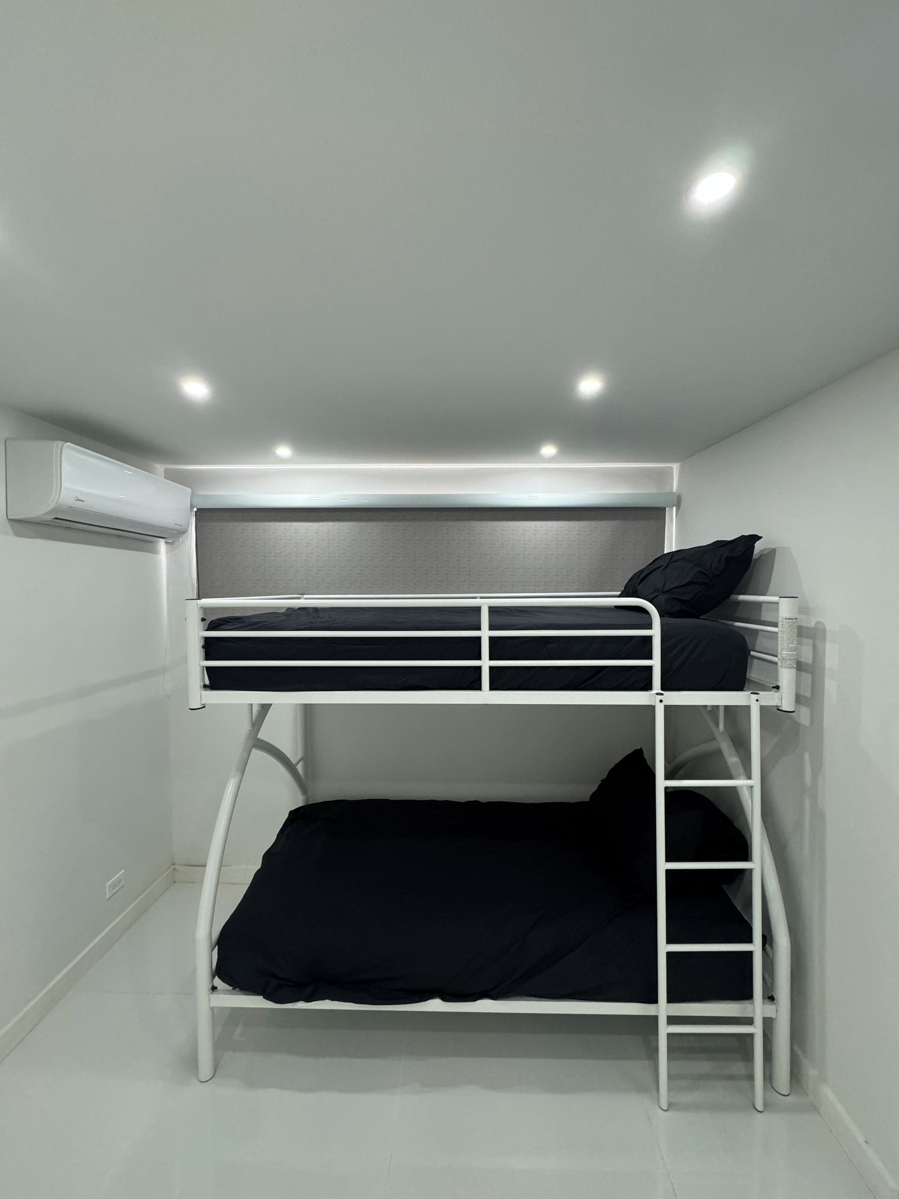 Cama individual con estructura blanca, cabecera gris y colchón negro. Hay un equipo de aire acondicionado blanco montado en la pared sobre la cama.