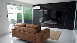 Uma sala de estar moderna com um sofá de couro marrom, uma porta de vidro que dá para uma área externa e uma parede revestida de azulejos pretos com uma televisão de tela plana.