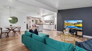 A modern living room with a green sofa, a dining area with a table and chairs, a kitchen with white cabinets, and a TV on the wall. - Spacious 5-bedroom POOL House with WiFi and AC in charming Chula Vista (Chula Vista)