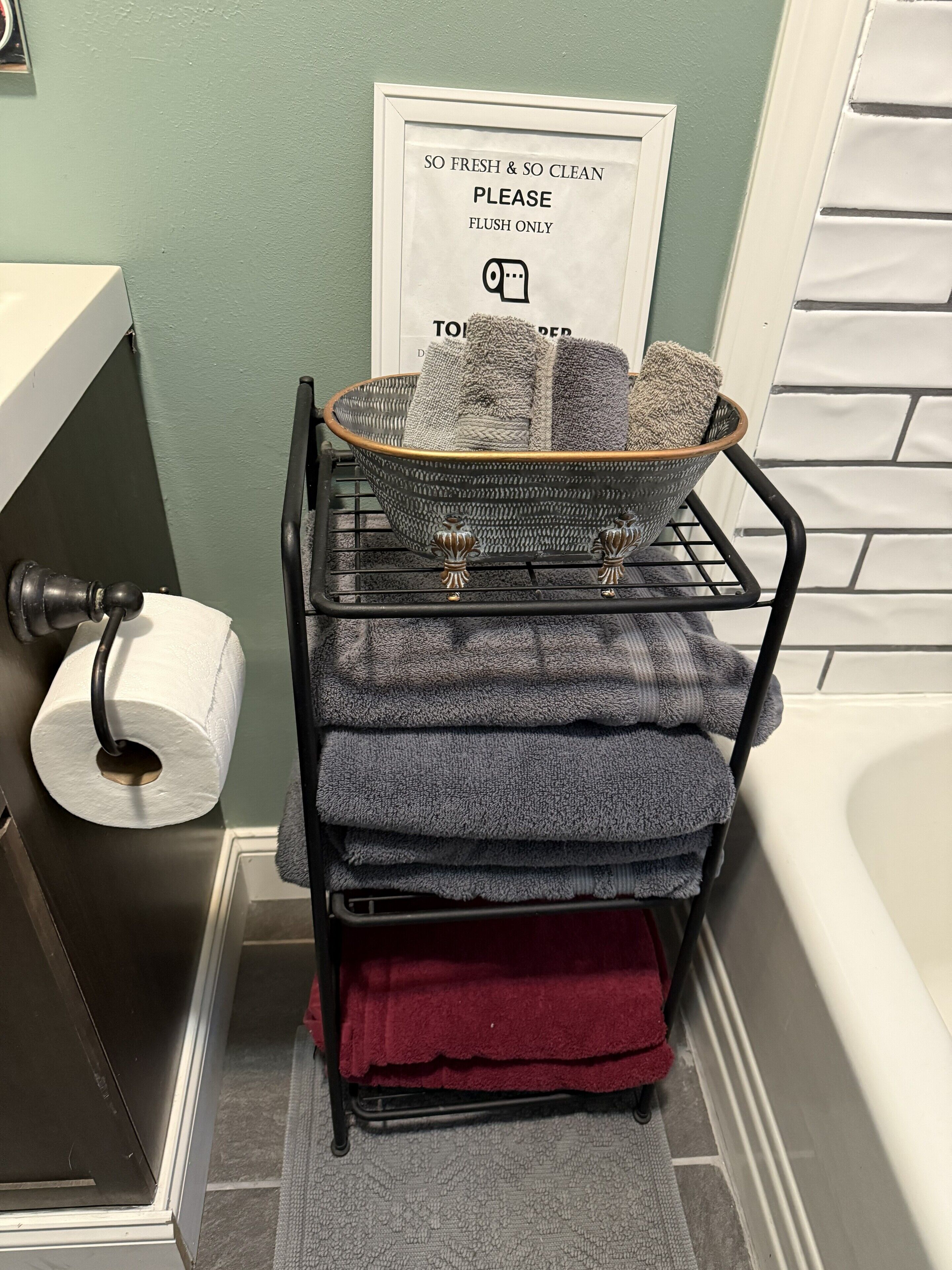 A bathroom with a black metal rack holding towels and a basket, a toilet paper roll, and a framed sign above the sink.