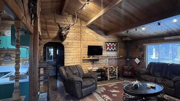 A log cabin interior with a fireplace, a mounted deer head, a television, and a cozy seating area.