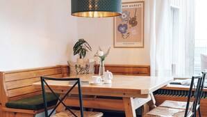 Une table à manger en bois, accompagnée d’un banc et de deux chaises à assise tressée, un luminaire suspendu, une plante en pot et un tableau encadré accroché au mur.