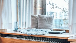Un coin salon confortable avec un banc rembourré, des coussins et un tapis moelleux, situé près d’une fenêtre dotée de stores verticaux.