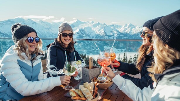 Un groupe de personnes trinquant avec des boissons autour d’une table en bois, avec une vue sur les montagnes en arrière-plan.