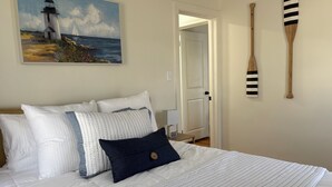 Una cama bien hecha con ropa de cama blanca y una almohada azul oscuro, un cuadro de un faro y dos remos de madera en la pared.