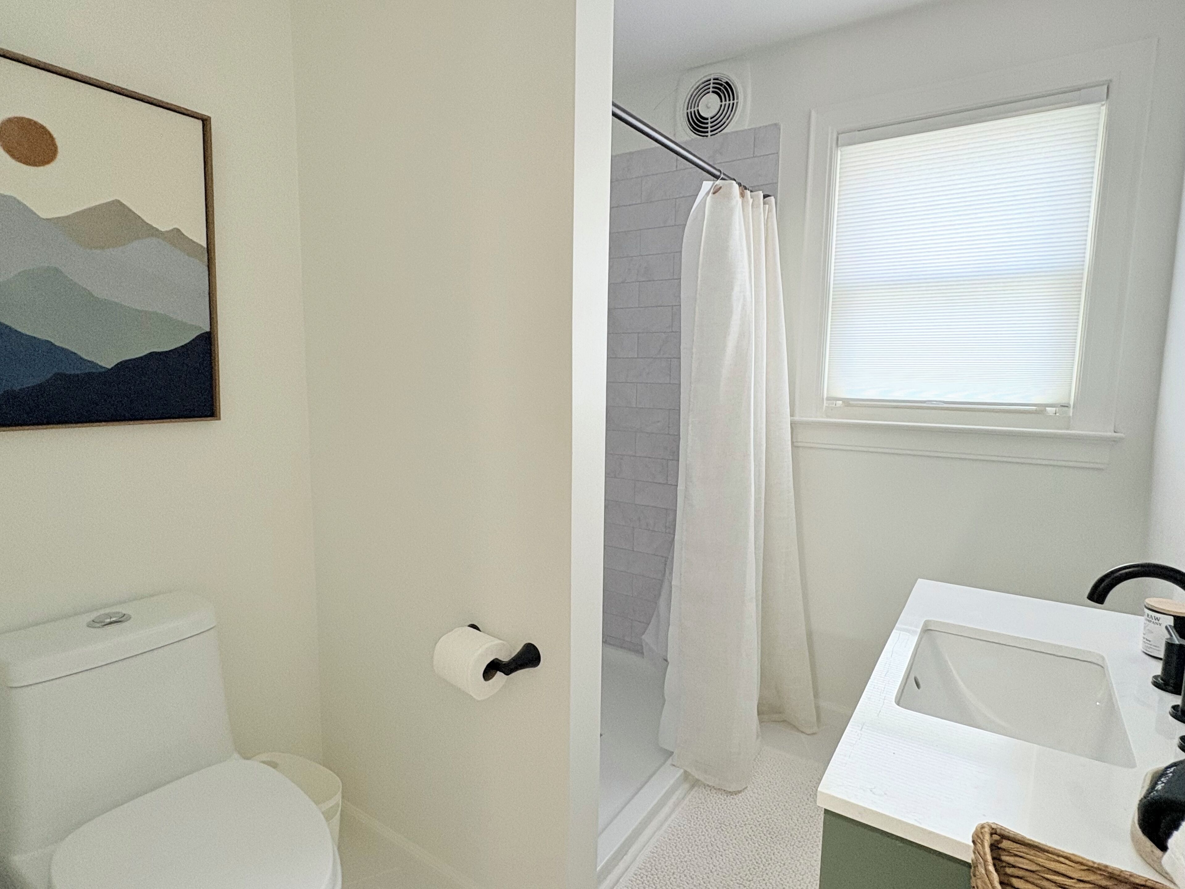 A bathroom with a toilet, a shower with a curtain, a sink, and a window with blinds.