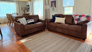 Una sala de estar con dos sofás de cuero marrón, un tapete estampado, suelo de madera, una zona de comedor con sillas y una mesa, y un ventanal con persianas.