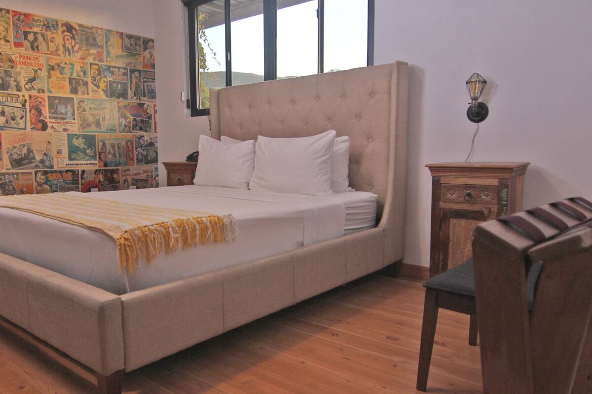 Une chambre avec un grand lit, un plancher en bois, un mur recouvert de vieilles affiches, une fenêtre et une table de chevet avec une lampe.