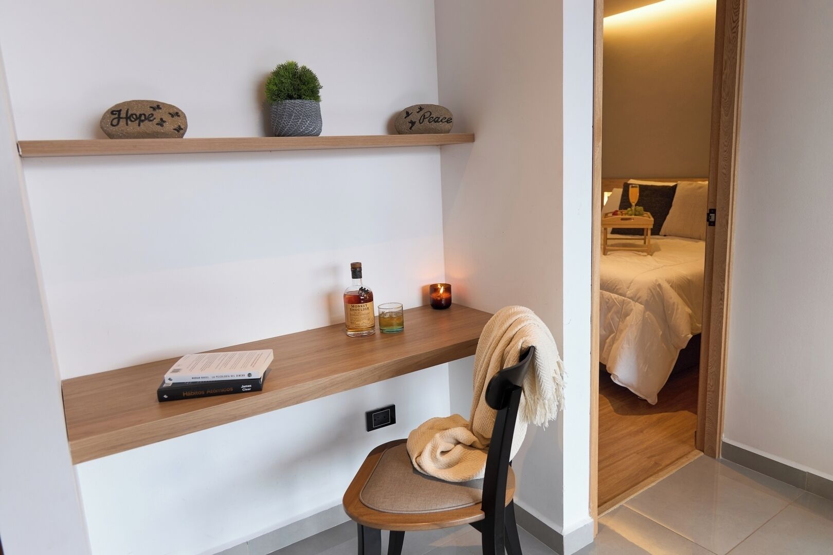 A modern room with a wooden desk, a chair, a bookshelf with decorative items, a bed with a white duvet, and a bedside table with a candle.