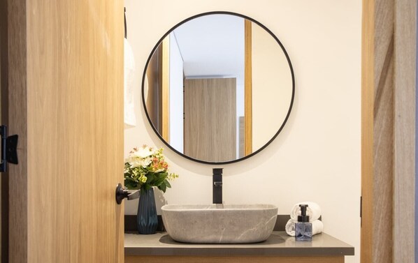 A bathroom with a round mirror, a vessel sink, a vase with flowers, and a towel dispenser. - Cozy Getaway Rosa Negra Stadio Suite (Medellín)