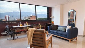 A modern living room with a blue sofa, a wooden chair, a dining table with chairs, and a large window offering a city view. - Elegant Cozy Suite Rosa Negra Stadio (Medellín)