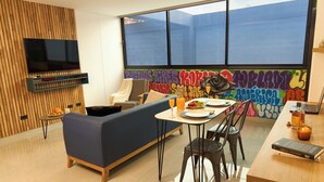A modern living room with a sofa, dining table, and chairs, a TV mounted on a wooden wall, and a colorful graffiti mural. - Charming Aparta-suite Stadio Rosa Negra Retreat (Medellín)