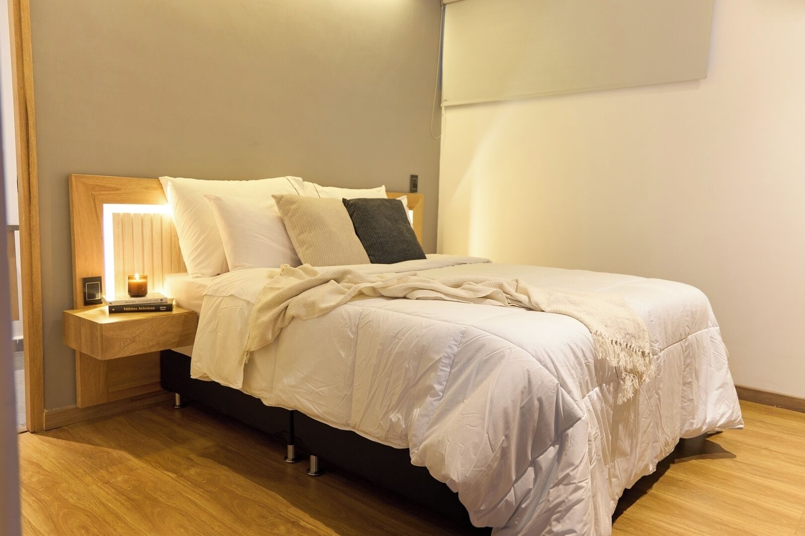 A neatly made bed with a white duvet, a bedside lamp, and a small table with a book and a candle.