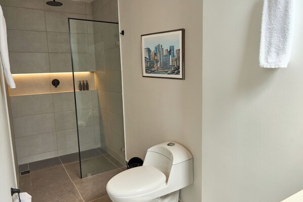 A modern bathroom with a glass-enclosed shower, a white toilet, and a framed cityscape picture on the wall. - Comfortable Escape Rosa Negra Stadio Apartasuite (Medellín)