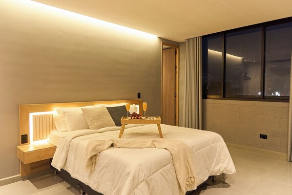 A modern hotel room with a large bed, bedside tables, a nightstand, and a view of the city at night. - Comfortable Escape Rosa Negra Stadio Apartasuite (Medellín)