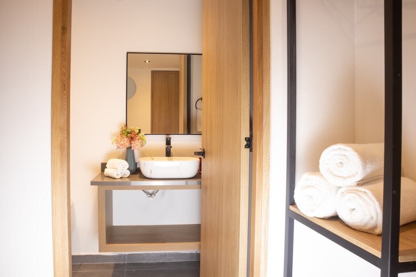 A bathroom with a wooden vanity, a white sink, and a mirror. There are towels and a vase with flowers on the vanity.