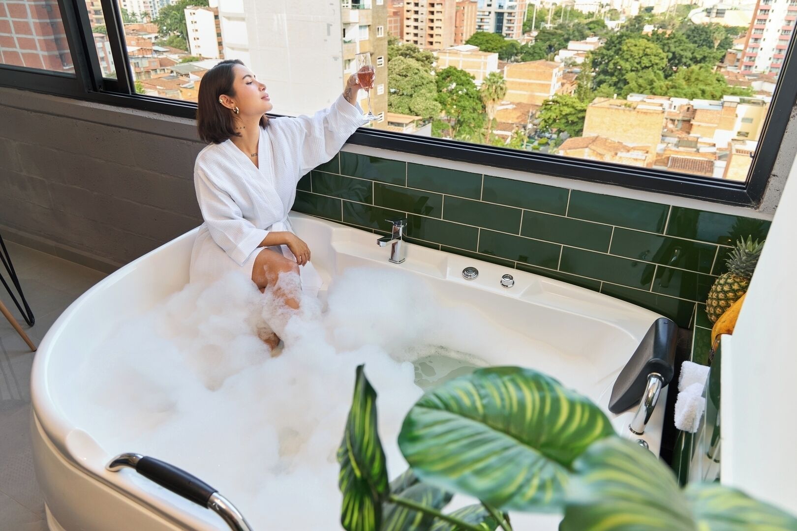 A person relaxing in a bathtub filled with bubbles, with a cityscape view through a window.
